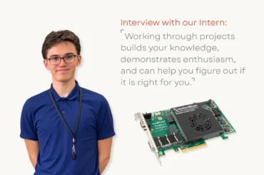 A student smiling at the camera next to a coaxpress over fiber frame grabber
